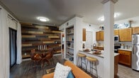 This well-appointed single-floor residence combines thoughtful design with eclectic character. The open-concept living and dining area immediately draws the eye with a striking reclaimed wood accent wall, rich dark hardwood-style flooring, and crown molding details. Multiple windows dressed with navy curtains and blinds fill the space with natural light while offering views of lush outdoor greenery. A built-in shelving unit adds both function and visual interest between the living and kitchen zones.

The kitchen is a functional galley-style layout outfitted with warm honey-toned cabinetry, granite-look countertops, a classic subway tile backsplash, and stainless steel appliances. A peninsula with bar-height seating bridges the kitchen to the living area, creating an ideal space for casual dining or entertaining.

The two bedrooms are carpeted and cozy, each with ceiling fans and windows that bring in natural light. One bedroom features a bold burnt-orange accent wall, adding a vibrant focal point to the room.

The bathroom is finished with wood-toned cabinetry, a granite-style vanity countertop, and warm overhead lighting, complemented by soft blue wall tones that lend a calm, cohesive atmosphere to the space overall.
