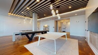 Located at 4011 Tryon Park Rd in Charlotte, NC, this contemporary amenity lounge delivers a refined social and recreational experience through thoughtful design and high-end finishes. Rich hardwood flooring flows throughout the open-plan space, anchoring the various activity zones and lending warmth to the modern aesthetic.

The games area serves as a central focal point, featuring a striking black shuffleboard table and a sleek white ping-pong table positioned atop a textured area rug. Overhead, a bold slatted wood ceiling treatment with integrated recessed lighting and cascading glass pendant fixtures adds dramatic visual depth. White textured tile accent walls provide a clean contrast to the warm wood tones throughout.

Along the perimeter, floor-to-ceiling windows flood the space with abundant natural light and offer open views of the surrounding landscape. Velvet bar-height chairs pair with pedestal tables to create an inviting lounge area ideal for casual gatherings. Sheer drapery softens the expansive glazing while maintaining the airy atmosphere.

Wide, art-lined corridors connect the main lounge to additional rooms, showcasing a curated collection of contemporary artwork and sculptural pieces that reinforce the space's gallery-like character. The overall ambiance is polished and welcoming, balancing modern architectural design with a relaxed, community-focused environment.