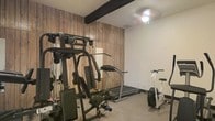 This single-floor fitness space offers a functional workout environment equipped with a variety of exercise equipment. The room features a multi-station cable gym machine as its centerpiece, complemented by a treadmill and a stationary bike, providing options for both strength training and cardiovascular exercise. A distinctive rustic wood-paneled accent wall adds warmth and character to the space, contrasting with the clean white walls on the adjacent side. An exposed dark structural beam runs across the ceiling, contributing an industrial aesthetic element. The room is lit by a ceiling-mounted light fixture, and the floor is covered in durable carpet suitable for a fitness environment. The layout is practical and efficient, with equipment arranged to allow reasonable circulation and use of each machine. The textured ceiling and neutral tones of the white walls create a balanced atmosphere that feels utilitarian yet inviting for exercise.