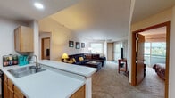 This single-floor residence offers a comfortable and functional layout designed for everyday living. The open-concept living room serves as the heart of the home, featuring vaulted ceilings that lend an airy sense of space, soft carpeting underfoot, and a sliding glass door that invites natural light and provides access to an outdoor balcony. The room flows seamlessly from the kitchen, which is separated by a breakfast bar with a white countertop. The galley-style kitchen is outfitted with warm oak cabinetry, black appliances including a range, microwave, and refrigerator, and tile flooring that adds a practical, easy-to-maintain surface. The two bedrooms are generously sized, each featuring plush carpeting, large windows for natural light, and ample space for full furnishings. Both bathrooms are finished consistently with warm oak vanities, white countertops, tile flooring, and tub-shower combinations with curtain enclosures. One bathroom features a well-lit vanity mirror with overhead lighting, while the other is equipped with a grab bar for added accessibility. Throughout the residence, warm wood-tone trim and neutral wall colors create a cohesive, welcoming ambiance that feels both practical and inviting.
