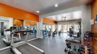 Located at 5104 E Van Buren St in Phoenix, Arizona, this energetic fitness and community center spans a generous single-floor footprint, thoughtfully divided into two distinct yet connected zones. The fitness area is boldly designed with striking orange accent walls, floor-to-ceiling mirrors, and durable dark rubber flooring. It is well-equipped with cardio machines including treadmills and ellipticals, strength training equipment such as a cable machine and weight rack, and a variety of free weights, offering a comprehensive workout experience. Recessed lighting and a ceiling fan ensure a comfortable environment throughout. French doors along the perimeter invite natural light and provide views of the outdoor surroundings. Transitioning into the adjacent multipurpose lounge area, the atmosphere shifts to a lighter, more relaxed tone. Light wood-look flooring, soft blue-gray walls, and recessed lighting create a welcoming, open setting. A long wall-mounted counter with colorful orange and yellow metal bar stools serves as a casual workspace or social gathering spot. Playful pop-art style wall décor adds personality to the space. This area also benefits from French door access to the outdoors, further connecting the interior to the surrounding community environment.