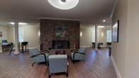 Located at 146 Yester Oaks Dr in Mobile, Alabama, this single-floor community space is thoughtfully designed to accommodate a variety of activities and social interactions. The open floor plan is unified by warm hardwood-style flooring, crown molding, and neutral taupe walls that create a welcoming, cohesive ambiance throughout.

A striking floor-to-ceiling brick fireplace serves as the centerpiece of the main lounge area, flanked by teal upholstered armchairs arranged to encourage conversation. Pendant drum lighting and recessed ceiling lights provide layered illumination across the expansive open space. Decorative vintage automobile-themed artwork adds a distinctive, lively character to the walls.

A dedicated media area features a large wall-mounted flat-screen television and a plush leather reclining sectional sofa set atop a vibrant multicolored area rug, offering a comfortable retreat for screen viewing. A separate, quieter seating nook with additional teal chairs and a sideboard provides a relaxed area for more intimate gatherings.

Towards one end of the space, a computer workstation area with multiple desks and chairs offers functional utility for residents or visitors. Elegant white columns punctuate the open layout, adding an architectural refinement. French doors lead to an adjoining room, enhancing the sense of connectivity and flow throughout this versatile, well-appointed community space.