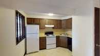 This single-floor unit offers a practical and comfortable layout with several notable features throughout. The kitchen is equipped with dark wood cabinetry providing ample upper and lower storage, yellow laminate countertops, a white electric range, a full-size white refrigerator, and a black dishwasher. A tile backsplash and patterned vinyl flooring complete the functional cooking space. A pass-through window connects the kitchen to the living area, enhancing the open feel between rooms.

The living area is generously sized, featuring neutral carpet and a textured ceiling. A standout architectural detail is a partial brick wall with decorative wooden pillars, adding character to the space. A sliding glass door floods the room with natural light and provides access to a private balcony with outdoor views.

Two carpeted bedrooms each include windows with blinds, offering natural light and a quiet atmosphere. Both rooms feature textured ceilings and neutral wall tones, providing a clean, move-in ready canvas.

The bathroom includes a full tub-shower combo with grab bars, a vanity with storage, a toilet, and a patterned tile floor. A hallway closet adjacent to the bathroom adds convenient storage. Overall, the unit has a classic, understated character with practical amenities throughout.