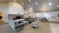 This well-appointed single-floor space offers an open, welcoming layout designed for comfort and community. The centerpiece of the main living area is a floor-to-ceiling stacked stone fireplace, framed by a crisp white surround that draws the eye upward toward the soaring vaulted ceiling. Recessed lighting throughout ensures the space remains bright and inviting, complementing the warm, neutral tones of the plush carpeted flooring. The seating area is generously furnished with plush sofas and accent chairs arranged around a sleek glass-top coffee table, creating an ideal environment for relaxed conversation and gathering. The open-plan layout flows seamlessly into a dining area at the rear, where a round dining table and chairs sit beneath elegant pendant light fixtures. Frosted glass panels and interior windows allow soft, diffused light to filter through from adjoining rooms or corridors, adding depth to the space while maintaining a sense of privacy. The overall ambiance is calm and refined, blending residential comfort with a functional, communal design — making it well-suited as a lounge, common area, or gathering room for residents or guests.