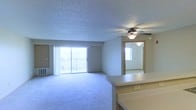 This single-floor unit presents an open-concept living area anchored by soft, neutral wall-to-wall carpeting that extends throughout the main space. A ceiling fan with an integrated light fixture is centrally mounted beneath a textured popcorn ceiling, providing both airflow and ambient lighting. Large sliding glass doors lead to a private balcony, allowing natural light to flood the interior and offering outdoor views. Vertical blinds frame the sliding doors, providing privacy and light control.

A built-in countertop or breakfast bar is visible in the foreground, hinting at a kitchen or dining area nearby, and features a conveniently located electrical outlet. The living area flows naturally toward an adjoining room, visible through a doorway with warm wood trim, which appears to be carpeted in a similar style and is lit by its own ceiling fixture. A wall-mounted heating or cooling unit is positioned near the balcony doors, ensuring year-round climate comfort. The overall layout is open and airy, making efficient use of the available space while maintaining a clean, functional aesthetic.
