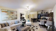This single-floor residence showcases an open-concept floor plan that seamlessly connects the living, dining, and kitchen areas into one cohesive and functional space. The living area is anchored by plush seating, a bold geometric area rug, and tasteful wall art, all set beneath a ceiling fan with integrated lighting. Neutral tones throughout the space create a warm and inviting ambiance, complemented by light-colored walls and carpeted flooring. The dining area transitions naturally from the living space, featuring a dark-toned table and chair set that adds contrast and visual interest. Adjacent to the dining area, the kitchen is equipped with dark cabinetry, black appliances including a refrigerator and range, and light-colored countertops that balance the darker elements. The kitchen is designed for both practicality and style, with ample cabinetry providing generous storage. Ceiling fans are present throughout, promoting comfortable airflow across the open layout. Interior doors visible in the background suggest access to additional private areas of the residence. Overall, the space presents a well-coordinated, move-in-ready interior with a clean and contemporary aesthetic suited for everyday living.
