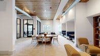 This thoughtfully designed single-floor communal space offers a generous open layout ideal for collaboration, relaxation, and focused work. The interior is anchored by striking wood-plank ceilings accented with exposed industrial-style ductwork, creating a warm yet contemporary aesthetic that blends modern and loft-inspired design elements.

The space is organized into several distinct functional zones. A central dining area features a large wooden table with mid-century modern chairs, perfect for group gatherings or shared meals. Along one wall, a series of semi-private navy blue upholstered booth seating nooks — each outfitted with a wooden table and pendant lighting — provide intimate spaces for smaller meetings or focused work sessions.

A relaxed lounge area offers dark leather sofas arranged around a coffee table beneath tall white walls and oversized windows that flood the room with natural light and offer views of the surrounding outdoor landscape. A cozy reading nook is anchored by a built-in wood bookshelf filled with curated books and décor, complemented by a pair of mustard-yellow lounge chairs. Black-framed glass partitions provide visual separation to an adjacent conference or office area. Soft carpet throughout ties the zones together, contributing to the space's welcoming and refined ambiance.