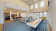 This single-floor amenity space is designed with a strong emphasis on social recreation and communal gathering. The game room is the centerpiece, featuring soaring double-height ceilings and an abundance of natural light streaming through a row of large arched windows positioned high along the upper walls, complemented by full-length French doors that open to outdoor views. The voluminous ceiling height creates an airy, grand atmosphere throughout. A pool table and shuffleboard table are the primary recreational fixtures, both finished in white with clean, modern lines and set atop a broad blue plaid-patterned carpet. Wall-mounted flat-screen televisions are positioned for optimal viewing. Adjacent seating areas with accent chairs provide comfortable spots to relax between games. The kitchenette area transitions seamlessly from the game room and features dark cabinetry contrasted by light quartz countertops, a herringbone brick tile backsplash, open display shelving, a stainless steel refrigerator, and a dedicated coffee station. Light wood-look flooring grounds the kitchen zone, while pendant lighting with gold accents adds a refined touch. Together, the spaces form a well-appointed, versatile environment suited for events and everyday recreational use.