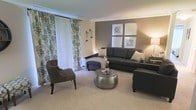 This single-floor space showcases a thoughtfully designed living area that balances comfort and style. The living room serves as the centerpiece, featuring a large, dark sectional sofa accented with patterned throw pillows, complemented by a plush upholstered armchair and matching ottoman. A distinctive hammered metallic round coffee table anchors the seating arrangement, adding a refined touch to the space. The walls are finished in a warm neutral palette, with a taupe accent wall serving as a sophisticated backdrop for a gallery arrangement of four framed artworks. Soft, wall-to-wall carpeting in a light beige tone runs throughout, contributing to the warm and inviting ambiance. Large windows dressed with elegant floral-patterned curtains in muted blue and cream tones allow generous natural light to filter into the room, enhancing the airy feel of the interior. Crown molding details add a polished architectural finish to the space. A floor lamp and wall-mounted sconces provide layered lighting options, creating a versatile atmosphere suited for both relaxed evenings and lively gatherings. Overall, this space presents a cohesive, well-curated interior that is both functional and welcoming.