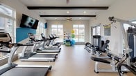 This fitness center offers a spacious, single-level layout designed to accommodate a comprehensive range of workout needs. The room is framed by large, multi-pane windows along multiple walls, flooding the space with natural light and providing views of the surrounding outdoor areas, including what appears to be a pool deck and landscaped grounds. Exposed dark wood ceiling beams add architectural character against the crisp white walls and ceiling, while recessed lighting and ceiling fans ensure comfort throughout the space.

The cardio section features a row of True-brand treadmills, upright and recumbent stationary bikes, and elliptical machines, all positioned to take advantage of the window views. A wall-mounted flat-screen television is positioned near the entrance for entertainment during workouts. The strength training area includes a multi-station cable machine, a full dumbbell rack with a wide range of weights, and adjustable benches. A dedicated functional training station holds stability balls and various accessories. The flooring throughout is a durable, light-toned exercise surface. Glass double doors provide easy access, contributing to an open, welcoming ambiance well-suited for daily fitness activity.