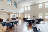Located at 6565 Chupp Rd in Stonecrest, GA, this single-floor community space is thoughtfully designed to accommodate gatherings and social interaction. The centerpiece of the space is a dramatic, double-height great room with soaring ceilings and rows of tall windows along multiple walls, flooding the interior with natural light and offering views of the surrounding greenery. Rich hardwood-style flooring runs throughout, providing a warm contrast to the crisp, neutral-toned walls.

The main gathering area is furnished with multiple round tables and contemporary upholstered chairs in deep navy blue, creating an inviting and flexible arrangement suited for group events or casual meetings. An upper-level mezzanine overlooks the main floor, adding architectural depth and visual interest to the space.

Adjacent to the great room is a well-appointed kitchen featuring dark espresso cabinetry, granite countertops with a hexagonal tile backsplash, stainless steel appliances, and a large island with bar seating — ideal for catering or communal use. A separate, more intimate lounge area offers a wall-mounted television, a plush sofa, and accent seating, providing a quieter retreat. Recessed lighting and ample window placement ensure the entire space remains bright and welcoming throughout.