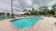 Located at 500 North Congress Avenue in Delray Beach, Florida, this outdoor pool area offers a classic South Florida resort-style experience. The centerpiece is a generously sized swimming pool with vivid turquoise water, clearly marked depth indicators, and a shallow wading area, making it suitable for a range of swimmers. The pool is bordered by a broad, interlocking paver deck that provides ample space for relaxation and movement around the water's edge.

Surrounding the pool, a collection of teal lounge chairs and tables are arranged beneath vibrant orange market umbrellas, offering shaded spots to unwind. The perimeter is enclosed by a white metal fence, ensuring safety while maintaining open sightlines to the lush greenery beyond. Towering palm trees and mature tropical foliage frame the space, creating a verdant, secluded atmosphere characteristic of South Florida's natural landscape. A multi-story residential building is visible in the background, contextualizing the pool as a shared community amenity.

The overall ambiance is relaxed and inviting, combining functional design with tropical aesthetics. The combination of the bright pool water, colorful furnishings, and surrounding palm trees creates a vibrant, resort-like setting well-suited for outdoor leisure in the warm Delray Beach climate.