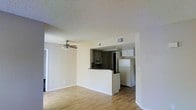 This single-floor residence offers a functional and open layout that seamlessly connects the living area to the kitchen. The living space features warm hardwood-style flooring that extends throughout, complemented by a ceiling fan that adds both practicality and a casual aesthetic. Neutral-toned walls keep the space feeling bright and versatile, while a textured popcorn ceiling adds a classic character overhead.

The kitchen is defined by a raised peninsula counter that serves as a natural divider between the cooking and living areas. Dark cabinetry lines the kitchen walls, providing ample storage, while essential appliances — including a refrigerator, microwave, and additional kitchen appliances — are neatly integrated into the space. The white refrigerator stands prominently at the kitchen's edge, easily accessible from the living area.

HVAC vents are visible in the ceiling, ensuring efficient climate control throughout the unit. A doorway off the living area hints at additional rooms beyond, suggesting a thoughtful and practical floor plan. Overall, the space presents a clean, straightforward layout suited for everyday living, with a cohesive design that balances utility and comfort.
