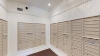 This dedicated mail room is a functional and neatly finished space designed to serve a large number of residents. The room is lined on multiple walls with banks of individual, labeled mailboxes finished in a neutral tan tone, providing organized and secure mail access for a substantial number of units. The mailbox units are recessed into the walls and include both standard-sized compartments and larger parcel lockers at the base, accommodating a variety of mail and package sizes.

The room features clean white walls accented by elegant crown molding that runs along the ceiling perimeter, lending a polished, classic aesthetic to what is otherwise a utilitarian space. Recessed ceiling lighting provides bright, even illumination throughout. The flooring consists of a durable, patterned carpet in muted tones, adding a touch of warmth and comfort underfoot. A security camera is mounted in the corner near the ceiling, ensuring the safety and security of residents' mail at all times. The overall design balances practicality with a refined finish consistent with a well-maintained residential building.
