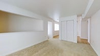 This single-floor residence presents a clean and functional layout designed for comfortable everyday living. The interior is finished with a neutral color palette throughout, featuring crisp white walls and ceilings that contribute to a bright, airy atmosphere. The main living area is finished with wall-to-wall beige carpet, providing a soft and warm underfoot experience. A distinctive built-in wall niche with a display shelf adds a subtle architectural detail, offering both visual interest and practical storage or display space. A set of bifold closet doors provides generous storage capacity, enhancing the room's overall utility. An open doorway leads to an adjoining room, which features hard surface flooring and a window that allows natural light to filter through, creating a seamless transition between spaces. Recessed ceiling vents are integrated throughout, ensuring consistent climate control. The overall design is straightforward and move-in ready, with an emphasis on clean lines, ample storage, and a welcoming, light-filled environment suited to a variety of living arrangements.