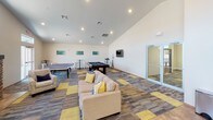 Located at 900 W Venture Pl in Sioux Falls, South Dakota, this single-floor community space is designed for social gathering and recreation. The open-concept layout is anchored by a generously sized lounge area featuring vaulted ceilings with recessed lighting, large clerestory windows that draw in natural light, and a striking stone fireplace with a classic mantel. Comfortable seating arranged around the fireplace creates a welcoming atmosphere for relaxed conversation or casual gatherings.

Adjacent to the lounge, a well-equipped kitchen features dark espresso cabinetry, granite-style countertops, full black appliances including a refrigerator and range, and a spacious island with bar-height seating, making it ideal for group events and catered functions. A dining area with tables and chairs provides additional seating nearby.

The game room area extends the space further, offering room for a full-size pool table, a ping pong table, and wall-mounted television. Patterned carpet tiles in blue, grey, and yellow add a contemporary visual element throughout, while glass doors provide access to an outdoor area. Together, these areas create a versatile, inviting environment suited for community use.