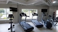 This fitness center offers a focused and functional workout environment designed to meet the needs of active users. The space is laid out on a single floor, providing a straightforward and accessible exercise area. The cardio zone is a highlight, featuring multiple treadmills and an elliptical machine positioned to take advantage of a large picture window that frames views of the surrounding greenery, filling the room with natural light and creating an open, energizing atmosphere. Wall-mounted flat-screen televisions are positioned on either side of the room, allowing users to stay entertained during their workouts. The interior maintains a clean, modern aesthetic with neutral gray walls, white-painted exposed ceiling beams, and a durable light-toned floor surface suited for high-traffic use. The layout provides ample spacing between equipment, ensuring comfort and safety during use. Free weights are also visible within the space, indicating a range of workout options beyond cardio. Overall, the fitness center strikes a balance between practicality and comfort, offering a bright, well-organized environment conducive to a productive exercise routine.