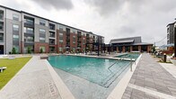 This expansive outdoor amenity space is the centerpiece of a modern multi-story residential community, showcasing thoughtfully designed features that blend style and functionality. The architecture of the surrounding buildings combines classic brick exteriors with contemporary gray paneling, large windows, and private balconies that overlook the courtyard below.

At the heart of the space is a generously sized lap pool with crystal-clear turquoise water, ideal for both recreation and fitness. Adjacent to the main pool is a dedicated spa or hot tub, offering a more intimate relaxation option. A separate shallow water feature with a surrounding fire pit and Adirondack-style seating provides a cozy gathering area, adding warmth and ambiance to the outdoor environment.

The sundeck is well-appointed with rows of lounge chairs and market umbrellas, offering ample space for residents to unwind. Sleek pergola structures with modern furnishings create shaded outdoor living areas, perfect for socializing. A dedicated clubhouse or amenity pavilion with a standing-seam metal roof anchors one end of the courtyard, providing additional covered space for gatherings.

Lush landscaping, including young trees and manicured greenery, softens the hardscape and adds a natural element throughout. The overall ambiance is polished, resort-inspired, and inviting, making this outdoor space a true community gathering hub.