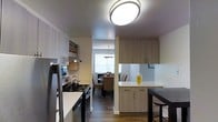 This single-floor residence offers a thoughtfully designed interior that blends functionality with modern style. The kitchen is a standout space, featuring sleek light-toned cabinetry that extends to the ceiling, providing generous storage throughout. Stainless steel appliances, including a refrigerator and a gas range, lend a contemporary feel, while a flush-mount circular ceiling light brightens the room with a clean, modern touch. A white countertop peninsula with additional cabinetry below serves as both a prep area and a casual dining spot, accompanied by bar-height seating. The open sightline from the kitchen leads directly into the dining area, which is illuminated by a decorative pendant light and benefits from natural light streaming through a window. The dining space comfortably accommodates a table with upholstered seating, creating an inviting area for meals and gatherings. Warm-toned wood-look flooring runs throughout, unifying the spaces and adding a sense of warmth. The overall ambiance is bright, clean, and contemporary, making excellent use of the available layout to create a cohesive and livable environment.