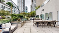 This well-appointed outdoor terrace offers a refined urban retreat nestled among the towers of a bustling city skyline. The space is thoughtfully organized into distinct zones, each serving a specific purpose while maintaining a cohesive aesthetic throughout. Large-format stone pavers define the primary walking and gathering surfaces, providing a clean and durable foundation across the entire terrace.

Multiple dining areas feature sturdy wooden tables surrounded by matching chairs, accommodating both intimate gatherings and larger group events. A dedicated lounge zone offers modular outdoor sofas and armchairs with plush white cushions, arranged around low coffee tables to encourage relaxed conversation. A compact putting green with artificial turf adds a recreational element, ideal for casual use and social engagement.

Glass-panel railings line the perimeter, preserving unobstructed sightlines to the lush garden beds below, which are planted with hydrangeas, ornamental grasses, ferns, and mature shrubs. These curated gardens provide a natural buffer and a touch of greenery against the surrounding urban backdrop. The terrace is bordered by the building's facade, featuring large sliding glass doors that connect the outdoor space directly to interior areas.

The overall ambiance balances urban sophistication with natural calm, offering an accessible and functional outdoor environment suitable for a variety of uses throughout the day.