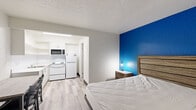 Located at 16410 North Fwy in Houston, TX, this efficiently designed single-floor unit offers a practical and well-appointed living space. The open-concept layout seamlessly integrates the sleeping area and kitchen, maximizing the use of available space. A bold blue textured accent wall serves as a standout design element in the main room, adding a pop of color against the neutral white and gray tones throughout.

The sleeping area features light wood-style laminate flooring, a wood-toned dresser, and a wall-mounted PTAC unit for climate control. A window with vertical blinds allows natural light to filter in, brightening the space. The kitchen is neatly tucked adjacent to the main room and includes white cabinetry, open upper shelving, a stainless steel sink, an electric range, a microwave, and a full-size refrigerator — providing all the essentials for in-unit cooking.

The bathroom is finished with tile flooring, a granite-style countertop vanity, a large wall mirror, recessed medicine cabinets, and a shower/tub combination accented by a navy and white striped curtain. Chrome fixtures and towel bars complete the clean, functional aesthetic. Overall, the unit offers a straightforward and efficient design well-suited for practical everyday living.