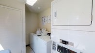 This small, single-floor laundry room is designed to maximize functionality within a compact footprint. The space features light-colored walls with a clean, utilitarian aesthetic, complemented by light tile flooring that contributes to an open, bright feel. Overhead, a flush-mounted fluorescent light fixture provides ample illumination throughout the room.

The laundry room is equipped with multiple top-loading washing machines positioned side by side along one wall, offering convenient access for users. A stacked dryer unit is situated near the entrance, making efficient use of vertical space to accommodate more machines within the limited floor area. Instructional and service information is posted on the wall near the washers, providing clear guidance for users on operating procedures.

A round dark accent rug near the entrance adds a practical touch, helping to manage foot traffic and maintain cleanliness. The room's straightforward, organized layout ensures ease of movement and access to all appliances. Overall, the space is a no-frills, well-equipped shared laundry facility designed with practicality and efficient use of space as its primary objectives.