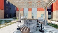 This single-floor unit opens to a well-appointed courtyard featuring a striking circular outdoor kitchen wrapped in stacked grey stone and topped with a granite-style counter. Two stainless steel grills flank the bar, which is surrounded by sleek adjustable bronze barstools. A white pergola overhead casts patterned shade across the concrete patio, creating an inviting al fresco entertaining space set against the building's bold red, orange, and tan exterior facade.

Inside, the kitchen is outfitted with rich dark espresso cabinetry paired with speckled granite countertops, providing a sharp contrast to the light-toned walls. Stainless steel appliances, including a dishwasher and range with overhead ventilation, complete the functional layout. The kitchen flows seamlessly into the open living area, where light hardwood-style flooring extends throughout and windows usher in generous natural light, creating a bright and airy atmosphere.

Interior doors feature a clean, paneled modern design with brushed nickel hardware, while louvered closet doors add a classic detail. The neutral white walls and light flooring throughout give the space a fresh, versatile canvas, while the overall layout balances open communal areas with clearly defined functional zones.