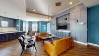 Located at 13444 Gran Bay Pkwy in Jacksonville, Florida, this single-floor community clubhouse is designed with a modern aesthetic that balances bold color and refined finishes. The lounge area serves as the heart of the space, featuring rich hardwood-style flooring, striking teal accent walls, and a paneled slate-blue feature wall that frames a wall-mounted flat-screen television above a built-in cabinet. Cognac leather sofas and industrial-style armchairs are arranged around a wood-and-metal coffee table, creating an inviting gathering environment. Overhead, a brass multi-arm chandelier adds a contemporary focal point, complemented by recessed lighting and sculptural swing-arm wall sconces. Elevated tray ceilings add architectural depth to the lounge. French doors along one wall open to an exterior area, allowing natural light to flow in and offering views to the outdoors. The kitchen area is equally well-appointed, featuring navy-blue cabinetry with a white subway tile backsplash, stainless steel appliances, and a generously sized island with a marble-look quartz countertop and bar seating. Pendant lighting above the island echoes the brass tones found throughout. Natural light enters through shuttered windows dressed with floor-length drape panels, enhancing the warm, welcoming ambiance of the entire space.