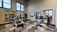 Located at 2817 Chad Dr in Eugene, Oregon, this contemporary fitness center is designed to support a full range of workout styles within a single, open-plan floor. The facility boasts soaring ceilings that lend an expansive, energetic atmosphere, complemented by large black-framed windows that flood the space with natural light and offer views of the surrounding community.

The flooring transitions seamlessly between two distinct zones: a bold hexagonal tile pattern anchors the cycling and stretching area, while a textured, geometric carpet defines the cardio and strength training sections. This thoughtful design detail helps visually organize the space without sacrificing openness.

Equipment is comprehensive and well-arranged. The cardio zone features treadmills and elliptical machines positioned to face the windows, alongside a row of Spinning bikes. The strength area includes a full dumbbell rack, adjustable benches, cable machines, and functional training rigs stocked with kettlebells, medicine balls, and resistance accessories. Wall-mounted mirrors amplify the sense of space and allow users to monitor their form throughout their workout.

A wall-mounted flat-screen display and motivational signage—including bold "Hustle, Grind, Execution" panels—reinforce an energizing atmosphere. An overhead ceiling fan and pendant lighting provide comfort and visibility. The space is accessible via glass-panel entry doors and includes clearly marked exits, making it both practical and welcoming for all fitness levels.
