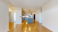 This single-floor residence is designed with a clean, contemporary aesthetic that emphasizes open living and functional design. The heart of the unit is the open-concept kitchen and living area, where light hardwood flooring flows seamlessly throughout. The kitchen is a standout feature, showcasing a substantial center island topped with striking blue-gray granite, complemented by a matching granite perimeter countertop. Gray shaker-style cabinetry with glass-front upper cabinets lines the walls, paired with a white subway tile backsplash and a full suite of stainless steel appliances, including a refrigerator with French doors, a gas range, and a built-in microwave. Three pendant lights with matte black hardware hang above the island, providing both task lighting and visual interest. The living area benefits from natural light streaming through French doors and a ceiling fan overhead. A dark barn-style door adds a modern-industrial accent near the entry. The bedrooms are carpeted for comfort, each featuring a ceiling fan and a window with exterior views. Hallways are finished in hardwood, maintaining continuity throughout. A spacious walk-in closet with wire shelving provides practical storage, and the overall white palette gives the unit a bright, move-in-ready feel.
