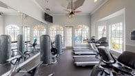 This fitness center offers a comprehensive workout environment designed with both functionality and aesthetic appeal in mind. The space is organized across a single floor and features distinct zones for cardio training and strength work, allowing for an efficient and varied exercise experience.

The cardio area is lined with multiple treadmills and elliptical machines, positioned to take full advantage of the abundant natural light streaming through large arched windows. These elegant windows, framed in white, offer views of outdoor greenery and lend the space a bright, airy quality uncommon in typical gym environments. Ceiling fans with decorative fixtures are mounted throughout, promoting comfortable air circulation during workouts.

The strength training area includes a selection of cable machines, weight stations, and a flat bench, accompanied by a full wall of mirrors that visually expand the space and allow users to monitor their form. Crown molding and white baseboards add a refined architectural touch, while the neutral gray walls and dark rubberized flooring provide a clean, modern contrast.

Overall, the fitness center balances practical workout amenities with thoughtful design details, creating an inviting and motivating environment for exercise.