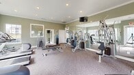 Located at 100 Belmere Luxury Ct in Houma, Louisiana, this fitness center offers a well-equipped and thoughtfully designed exercise room. The space is finished with soft sage-green walls, crown molding, and plush carpeted flooring, lending an upscale aesthetic that sets it apart from a typical gym environment. Recessed lighting throughout ensures bright, even illumination, while large windows along multiple walls flood the room with abundant natural light and provide views of the surrounding outdoor grounds. Full-length mirrors are strategically positioned to visually expand the space and allow users to monitor their form during workouts. The cardio section features multiple treadmills and an elliptical machine, all positioned to take advantage of the natural light and outdoor views. The strength training area is comprehensively stocked with a multi-station cable and weight machine, a free weight rack with a full dumbbell set, and various adjustable benches. A wall-mounted flat-screen television adds convenience for entertainment during workouts. A water cooler is conveniently available within the room. Access is provided through both a standard door and a glass-panel door, contributing to an open and airy feel throughout this polished, functional facility.