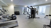 This fitness facility offers a thoughtfully organized workout space designed to accommodate a wide range of exercise routines. The room features durable, light-toned modular flooring that provides both comfort and stability underfoot, while a drop ceiling with recessed lighting ensures bright, even illumination throughout.

Strength training is well-represented with a multi-station cable machine that supports a variety of resistance exercises, a dedicated dumbbell rack stocked with multiple weight increments, and a weight bench for targeted lifts and presses. A large wall-mounted mirror enhances the sense of space and allows users to monitor their form during workouts.

Cardio equipment is also present, including a treadmill positioned near the entrance of the space, making it easy to warm up or cool down. The overall layout is open and uncluttered, allowing for free movement between stations.

The walls are finished in a neutral gray tone, contributing to a clean and focused atmosphere. Natural light filters in through a window, complementing the recessed ceiling lights to maintain a well-lit environment. The space is designed with functionality at its core, offering everything needed for a balanced and effective fitness regimen.