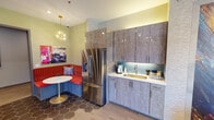 Located at 1701 Pharr St in Fort Worth, TX, this single-floor space showcases a bold, eclectic design aesthetic that blends color, texture, and contemporary furnishings throughout. The layout is thoughtfully organized, transitioning seamlessly between distinct functional areas connected by well-appointed corridors.

The kitchenette area is a standout feature, combining practicality with personality. Textured wood-grain cabinetry extends floor to ceiling, complemented by a teal hexagonal tile backsplash, white countertops, and a stainless steel refrigerator. A cozy built-in banquette with vibrant red upholstery and a round pedestal table occupies the corner, illuminated by a striking gold starburst pendant light. The flooring features large hexagonal tiles, adding a retro-modern touch.

The lounge area commands attention with a dramatic feature wall of richly colored abstract artwork serving as a backdrop for a wall-mounted flat-screen TV and a sleek linear fireplace below. Comfortable gray lounge chairs with orange accent pillows are arranged around a rustic drum coffee table atop a plush area rug, creating an inviting gathering space.

Connecting hallways are adorned with vibrant, curated artwork and recessed lighting, maintaining the space's energetic character while guiding visitors between areas. The overall ambiance is modern, artistic, and welcoming.