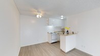 This 1-bedroom, 1-bathroom unit is laid out entirely on a single floor, offering a streamlined and functional living experience. The open-concept design connects the living area directly to the kitchen, creating a sense of openness and ease of movement throughout the space. Light-toned wood-style flooring runs throughout, contributing to a clean and airy aesthetic that reflects natural light effectively. The living area features a ceiling fan with integrated lighting, providing both comfort and illumination. The kitchen is outfitted with white cabinetry that extends to the upper walls, maximizing storage. A gas range and sink are neatly integrated into the layout, and a peninsula counter with a warm-toned surface serves as a practical divider between the kitchen and living area — ideal for casual dining or additional workspace. Open shelving above the kitchen counter adds further storage and display options. The overall color palette is bright and neutral, with white walls and ceilings — including a textured ceiling finish — creating a cohesive and welcoming ambiance throughout. The thoughtful layout makes efficient use of the available space, making it a well-suited option for individuals or couples seeking a comfortable and low-maintenance home.
