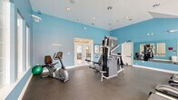 Located at 3250 Carter Rd in Sumter, South Carolina, this well-equipped fitness center offers a dedicated space for residents to maintain an active lifestyle. The room is defined by striking sky-blue walls, bright white trim, and a vaulted ceiling that lends a sense of openness and volume to the space. Recessed lighting supplements the abundant natural light that pours in through multiple large windows and a set of classic French doors, keeping the environment bright and energizing throughout the day. The durable rubberized flooring provides a safe, easy-to-clean surface suitable for a wide range of workouts. The fitness floor is outfitted with a comprehensive selection of equipment, including treadmills, elliptical cross-trainers, a recumbent stationary bike, and a multi-station cable strength machine, catering to both cardio and resistance training needs. A wall-mounted mirror along one side of the room enhances the sense of space and allows users to monitor their form during exercise. Workout reference posters are displayed on the walls, adding both a functional and decorative touch. An adjacent windowed room is visible from the main floor, and a wall-mounted television provides entertainment during workouts. The overall ambiance is clean, well-maintained, and purposefully designed to support an effective fitness experience.
