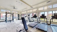 This fitness center is a thoughtfully designed, single-floor facility that offers a comprehensive workout environment for all fitness levels. The space is characterized by an abundance of natural light, courtesy of large floor-to-ceiling windows that line the walls and provide open views of the surrounding exterior. Upper transom windows further enhance the bright, airy atmosphere throughout the day.

The cardio zone is well-appointed with multiple treadmills, elliptical trainers, a stair climber, and a stationary spin bike — each positioned to take advantage of the outdoor views. A dedicated strength training area features a multi-functional cable and power rack system, a weight bench, and a full dumbbell rack, catering to a wide range of resistance training needs. Ceiling fans overhead provide consistent airflow and comfort during intense workouts.

The interior design is clean and contemporary, with patterned low-pile carpet flooring, recessed lighting, and a neutral warm-white color palette accented by dark charcoal architectural columns. Floor-to-ceiling mirrors are strategically placed along one wall, visually expanding the space and supporting proper form during exercise. Entry is provided through sleek glass-panel double doors. The overall ambiance is professional, energizing, and inviting.