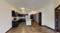 This well-appointed single-floor residence offers a thoughtfully designed layout centered around an open-concept kitchen and living area. The kitchen is a standout feature, showcasing rich dark cabinetry that contrasts elegantly with granite countertops and a central island with pendant lighting above. Stainless steel appliances, including a refrigerator, range, and microwave, provide a sleek, modern finish. Durable wood-look flooring runs throughout, adding warmth and cohesion to the space.

The layout extends naturally from the kitchen toward a hallway leading to additional rooms, with dark-stained doors that complement the overall color palette. Flush-mount ceiling fixtures provide consistent, ambient lighting throughout the main living area.

One of the most notable features of this residence is the private balcony, which offers serene, unobstructed views of lush green trees and a landscaped lawn below. The outdoor space provides a tranquil retreat with a wrought-iron railing, making it an ideal spot for relaxing while enjoying the natural surroundings. Together, the modern interior finishes and scenic outdoor views create a balanced and comfortable living environment.