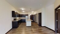 This well-appointed single-floor residence offers a thoughtfully designed layout centered around an open-concept kitchen and living area. The kitchen is a standout feature, showcasing rich dark cabinetry that contrasts elegantly with granite countertops and a central island with pendant lighting above. Stainless steel appliances, including a refrigerator, range, and microwave, provide a sleek, modern finish. Durable wood-look flooring runs throughout, adding warmth and cohesion to the space.

The layout extends naturally from the kitchen toward a hallway leading to additional rooms, with dark-stained doors that complement the overall color palette. Flush-mount ceiling fixtures provide consistent, ambient lighting throughout the main living area.

One of the most notable features of this residence is the private balcony, which offers serene, unobstructed views of lush green trees and a landscaped lawn below. The outdoor space provides a tranquil retreat with a wrought-iron railing, making it an ideal spot for relaxing while enjoying the natural surroundings. Together, the modern interior finishes and scenic outdoor views create a balanced and comfortable living environment.