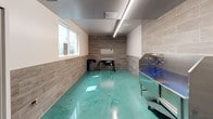 Wash & Wag is a professionally designed pet grooming facility built with both functionality and cleanliness in mind. The single-floor space is defined by its striking teal epoxy flooring, which offers a durable, easy-to-clean surface well-suited for a high-use grooming environment. The walls feature large-format wood-look ceramic tile wainscoting that extends halfway up, adding a warm, contemporary aesthetic while providing a practical, moisture-resistant surface.

The facility is equipped with stainless steel wash stations fitted with coiled blue hoses, designed for efficient and hygienic pet bathing. A dedicated grooming table with a stainless steel surface and integrated dryer provides a designated area for post-wash grooming and styling. Overhead, linear LED lighting runs the length of the ceiling, ensuring bright, even illumination throughout the workspace.

Natural light filters in through multiple windows, contributing to an open and well-lit atmosphere. Ceiling-mounted ventilation keeps the air fresh and the environment comfortable. Access doors at either end of the space provide smooth traffic flow during operations. The overall design balances a clean, clinical efficiency with a modern, welcoming aesthetic, making Wash & Wag a well-appointed destination for pet care.