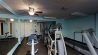 This single-floor fitness room offers a functional workout environment featuring an assortment of exercise equipment suited for both cardio and strength training. The space is painted in a light teal/blue tone, contributing to a calm and energizing atmosphere. Overhead, a ceiling fan with an integrated light fixture provides air circulation and ambient lighting, complemented by additional ceiling-mounted light fixtures throughout the room. The textured ceiling adds a classic architectural detail to the space. Equipment includes an elliptical trainer, a multi-station cable weight machine, a recumbent bike, a treadmill, and what appears to be an adjustable bench or incline trainer, providing users with diverse workout options. A fire extinguisher is mounted on the wall, reflecting attention to safety. The room also features a doorway leading to an adjacent area, suggesting access to additional amenities. Dark baseboards frame the perimeter of the room, providing a subtle contrast to the light-colored walls. The overall layout is efficiently organized to maximize the use of the available floor space.