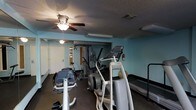 This single-floor fitness room offers a functional workout environment featuring an assortment of exercise equipment suited for both cardio and strength training. The space is painted in a light teal/blue tone, contributing to a calm and energizing atmosphere. Overhead, a ceiling fan with an integrated light fixture provides air circulation and ambient lighting, complemented by additional ceiling-mounted light fixtures throughout the room. The textured ceiling adds a classic architectural detail to the space. Equipment includes an elliptical trainer, a multi-station cable weight machine, a recumbent bike, a treadmill, and what appears to be an adjustable bench or incline trainer, providing users with diverse workout options. A fire extinguisher is mounted on the wall, reflecting attention to safety. The room also features a doorway leading to an adjacent area, suggesting access to additional amenities. Dark baseboards frame the perimeter of the room, providing a subtle contrast to the light-colored walls. The overall layout is efficiently organized to maximize the use of the available floor space.