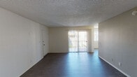 This single-floor unit offers a straightforward and functional layout across several well-defined spaces. The living area is generously proportioned, featuring dark hardwood-style flooring that contrasts cleanly with the neutral white walls and textured popcorn ceiling. A full-width sliding glass door dressed with vertical blinds fills the room with natural light and provides direct access to an outdoor area, while an additional window further brightens the space. A secondary room with light-toned wood-style flooring and warm beige accent walls offers a versatile space suitable for a bedroom or additional living area, with a partial dividing wall adding subtle definition to the layout. The kitchen is efficiently designed with ample white cabinetry extending to the ceiling for generous storage, granite-style countertops, a built-in dishwasher, an electric range with overhead ventilation, and a single-basin sink — all finished in a cohesive white palette with wood-style flooring underfoot. The bathroom features a wide vanity with granite-style countertop, an undermount sink, a large wall mirror, and a three-bulb vanity light fixture, with a combined tub and shower visible in the background. Natural light from a small window adds brightness to the space. Overall, the unit presents a clean, neutral, and move-in-ready environment.