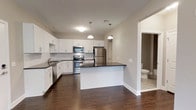 Located at 2000 Birchwood Circle in Bethlehem, Pennsylvania, this single-floor residence offers a thoughtfully designed open-concept layout that maximizes flow and functionality. The kitchen serves as the centerpiece of the space, showcasing crisp white shaker-style cabinetry, dark stone countertops, and a full suite of stainless steel appliances, including a range, microwave, and refrigerator. A central island with a matching dark countertop provides additional workspace and a natural gathering point. Pendant lighting fixtures hang above, adding both warmth and visual interest to the kitchen area. Rich, dark hardwood flooring runs throughout the main living area, creating a cohesive and elegant aesthetic that contrasts beautifully with the white cabinetry and neutral gray walls. The open layout allows the kitchen to connect seamlessly to the adjacent living space, enhancing the sense of spaciousness. A bathroom is conveniently accessible just off the main living area, featuring tile flooring. The overall ambiance is clean, contemporary, and move-in ready, with a neutral palette that provides a versatile backdrop for a variety of interior styles.