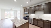 This single-story residence at 15840 Championship Pkwy in Fort Worth, Texas, offers a thoughtfully designed interior with modern finishes throughout. The kitchen serves as the centerpiece of the home, showcasing rich dark-toned cabinetry, gleaming granite countertops with subtle veining, and a full suite of stainless steel appliances including a range, microwave, dishwasher, and refrigerator. A classic white subway tile backsplash adds texture and contrast, while a central island provides additional prep space and casual seating. Light wood-tone flooring runs throughout the main living areas, lending warmth and continuity to the open layout. Recessed lighting and a ceiling fan ensure the space is both well-lit and comfortable. The bathroom features a clean, minimalist design with a full soaking tub and shower combination, accented by white stacked tile walls and light wood-style flooring. The bedroom is carpeted for comfort and benefits from a large window that frames views of the surrounding community green space, filling the room with natural light. The overall aesthetic is contemporary and cohesive, combining neutral tones, quality materials, and functional design elements throughout.