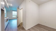 This single-floor unit presents a clean, contemporary design highlighted by neutral white walls, white trim, and wood-look plank flooring that flows consistently throughout the space. The layout is efficiently organized, connecting a main living area, a kitchenette, a bathroom, and a separate room accessible through an interior door.

The living area is bright and open, anchored by a large picture window that frames an elevated urban view of the surrounding streetscape and neighborhood. A ceiling fan provides both light and air circulation in this room. The elevated position of the unit allows for an expansive outlook, adding a sense of openness to the interior.

The kitchen features white cabinetry with brushed nickel hardware, a smooth white countertop, an integrated cooktop, a single-basin sink with a modern gooseneck faucet, and open upper shelving alongside upper cabinets with a ventilation hood — all finished in a cohesive, minimalist palette with light blue accent walls.

The bathroom is thoughtfully appointed with a wall-mounted white vanity, an undermount sink, a large frameless mirror, and matching wood-look flooring, maintaining the unit's consistent modern aesthetic. A floor-level vent and recessed sprinkler heads are noted throughout, reflecting practical building infrastructure integrated neatly into the design.