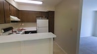 This single-floor space includes a kitchen that serves as a central feature, designed with practicality in mind. The kitchen is outfitted with dark wood cabinetry that extends to the upper walls, providing substantial storage space. White appliances, including a full-size refrigerator and a range with a black dishwasher, are integrated into the layout, offering a contrast against the darker cabinetry. A white peninsula extends from the kitchen, creating a defined boundary between the kitchen and the adjacent area, while also functioning as a convenient workspace or casual dining surface. Overhead, a fluorescent light fixture provides direct illumination across the kitchen workspace. The countertops are finished in white laminate, maintaining a clean and cohesive aesthetic. Neutral-toned walls and light carpeting in the adjacent area contribute to a simple, understated ambiance throughout the space. The overall layout is compact yet organized, making efficient use of the available space while keeping functionality as a priority.