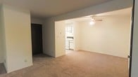 This single-floor space features an open-concept layout that connects the living area to the kitchen, creating a functional and cohesive flow throughout. The spacious living area is finished with wall-to-wall carpet flooring in a neutral tone, lending a comfortable and understated feel to the room. A ceiling fan with a light fixture is mounted overhead, providing both air circulation and ambient lighting. The walls are painted in a light, neutral color throughout, contributing to a clean and bright atmosphere.

The kitchen is visible through an open passageway and is outfitted with white cabinetry, white appliances including a range and dishwasher, and light-colored countertops. The kitchen's bright finish complements the overall neutral palette of the space. A doorway on the left side of the living area leads to an additional room, suggesting further private spaces beyond the main open area. The straightforward, functional design of the unit makes it a versatile space well-suited for a variety of living arrangements.