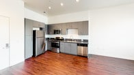 Located at 3050 Mission Inn Ave in Riverside, California, this single-floor unit offers a thoughtfully designed layout with modern finishes throughout. The kitchen is a standout feature, showcasing sleek gray flat-front cabinetry extending to the ceiling for ample storage, dark countertops, and a full suite of stainless steel appliances including a refrigerator, electric range, over-the-range microwave, and dishwasher. Recessed lighting illuminates the space, while rich hardwood-style flooring ties the kitchen and open living area together seamlessly. The bedroom is generously proportioned, featuring soft carpeting, crisp white walls, and a large window that invites abundant natural light and views of the surrounding neighborhood. The vaulted ceiling adds a sense of openness and volume to the room. The bathroom is clean and functional, finished with hardwood-style flooring consistent with the rest of the unit, a gray vanity with a built-in sink, a large mirror, and a full tub-shower combination with white tile surround. The overall aesthetic of the unit is contemporary and understated, with a neutral palette that provides a versatile backdrop for a variety of interior styles.