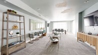 Located at 401 N Chaparral St in Corpus Christi, Texas, this single-floor multipurpose room serves as both a professional conference space and a relaxed lounge area. The open layout is unified by patterned, light-gray carpeting laid in a herringbone design, which adds visual texture and warmth throughout the space.

The conference area is anchored by a large rectangular wood-toned table surrounded by ergonomic rolling chairs, accommodating sizable group meetings. A wall-mounted flat-screen TV is positioned nearby for presentations and collaborative work. Sheer white floor-to-ceiling drapes line the exterior wall, diffusing generous natural light and contributing to the room's airy, calm atmosphere.

The lounge area features a curated mix of modern accent chairs with gold-toned frames, a small side table, and additional seating options including a warm leather chair near the entry. A decorative open shelving unit and a wooden sideboard provide functional display surfaces, while framed artwork and a table lamp add refined finishing touches.

The soft blue-gray walls create a cohesive, professional aesthetic throughout the space. Glass double doors at the entry provide easy access and a transparent connection to the adjacent corridor. Overall, the space is well-suited for meetings, community gatherings, or collaborative work sessions.